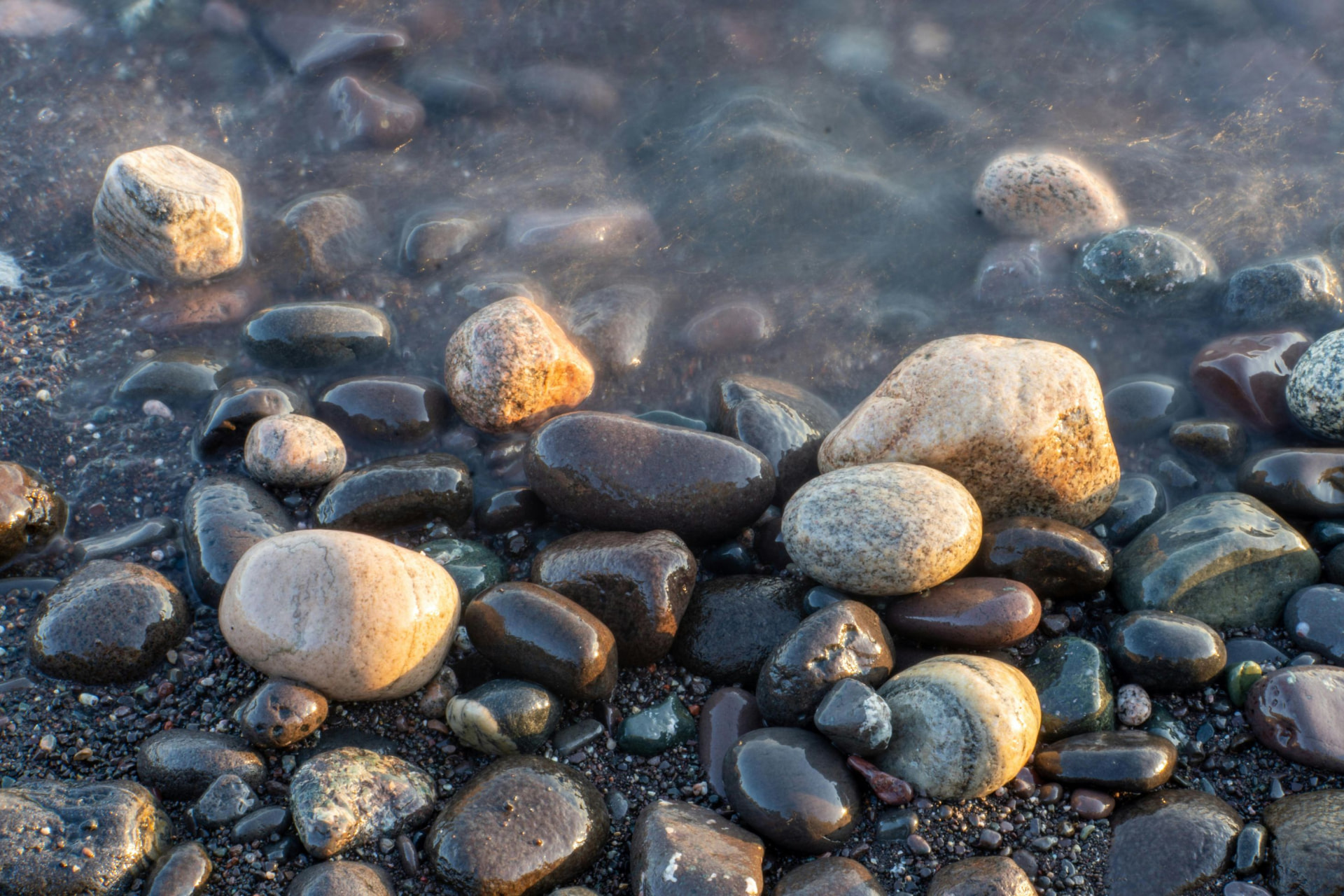 Smooth rounded stones in earthy tones along the Lake Michigan shoreline, with soft water washing over them at the water's edge